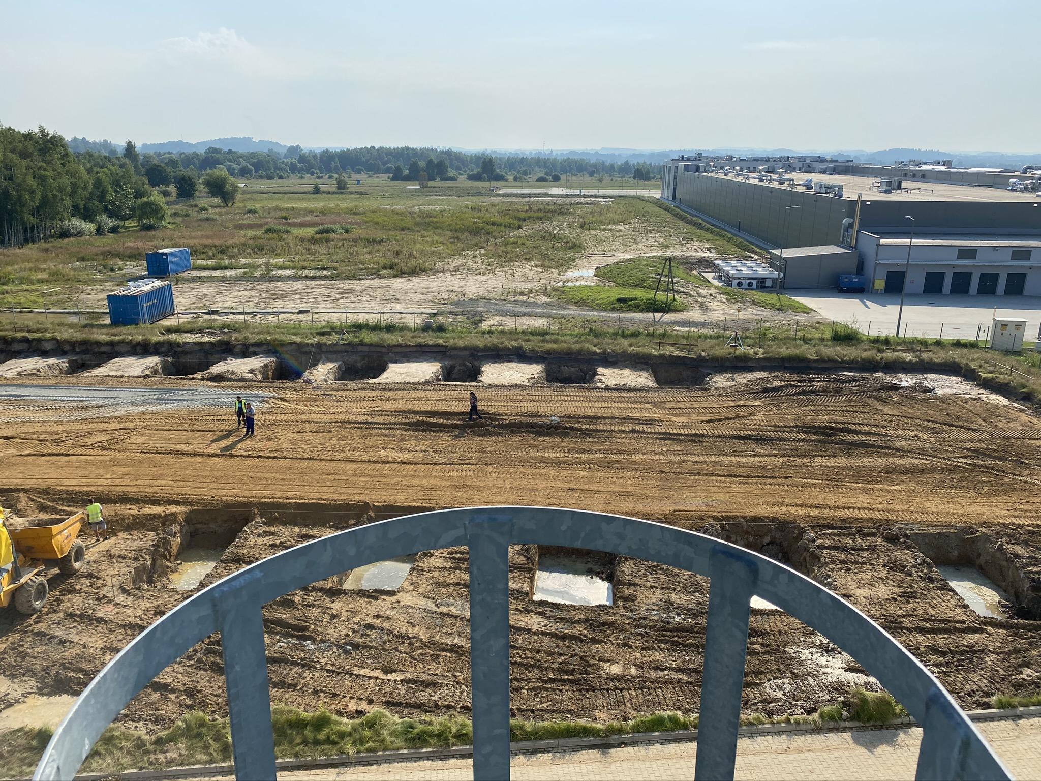 Szeroki widok z góry na plac budowy. W centrum widać świeżo przekopaną, brązową ziemię z rzędami prostokątnych wykopów pod fundamenty. W oddali po prawej stronie znajduje się duży, długi budynek magazynowy, a reszta otoczenia to zielone, nieużytkowe tereny. Na pierwszym planie znajduje się fragment stalowej konstrukcji, prawdopodobnie poręczy.
