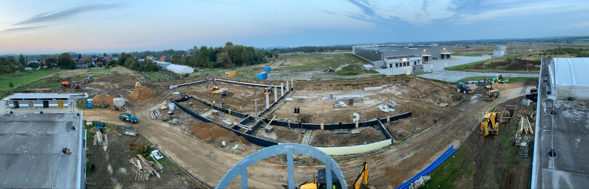 Panoramiczny widok z góry na duży plac budowy. W centrum zdjęcia widać świeżo wykonane fundamenty budynku o zakrzywionym kształcie, z licznymi, pionowymi słupami nośnymi. Wokół fundamentów rozciąga się nierówny teren z drogami gruntowymi i sprzętem budowlanym. W tle widoczne są zabudowania mieszkalne i tereny zielone, a po bokach fragmenty istniejących budynków magazynowych.