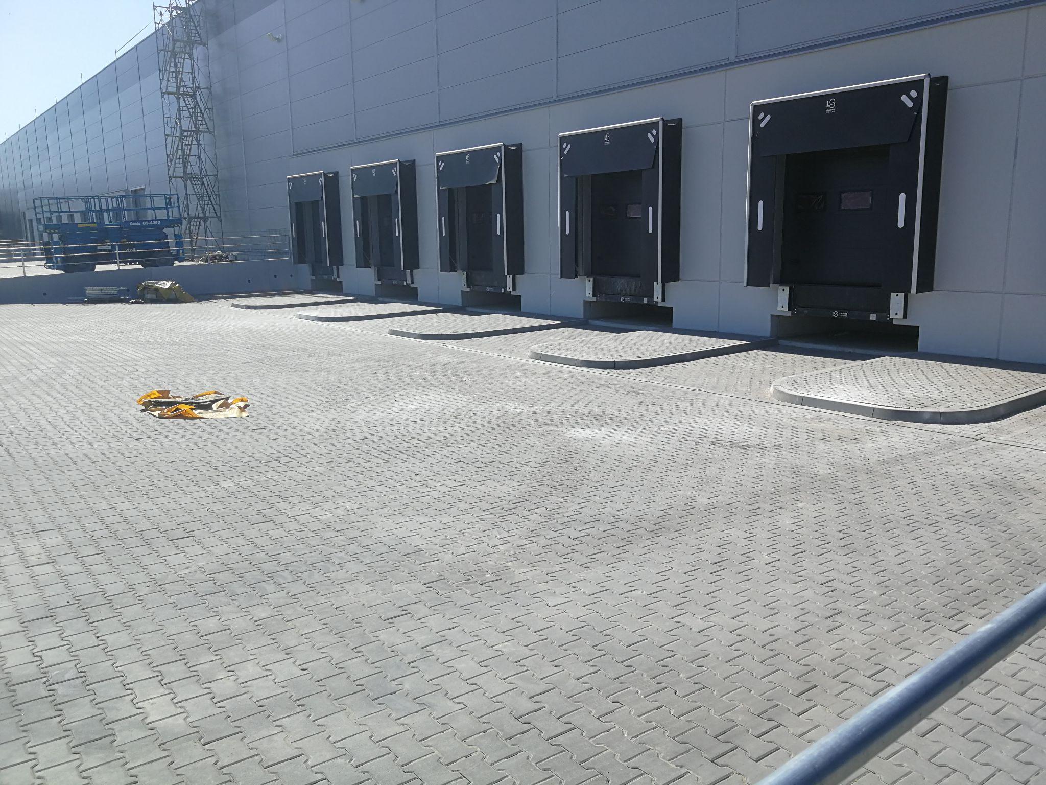 Zewnętrzna ściana nowoczesnego magazynu logistycznego z pięcioma rzędami doków przeładunkowych. Doki mają czarne, gumowe uszczelnienia. Przed nimi rozciąga się duży, wybrukowany plac manewrowy. Po lewej stronie widać rusztowanie i podnośnik nożycowy.