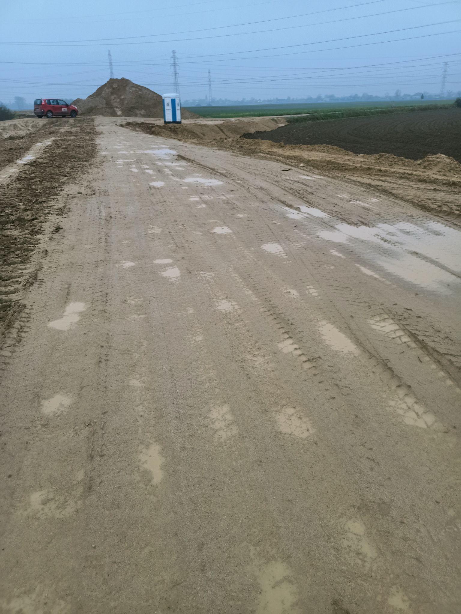 Droga gruntowa na placu budowy po deszczu. Nawierzchnia jest piaszczysta i błotnista, z widocznymi śladami opon i kałużami. Po bokach drogi widać nasypy z ziemi. W oddali po prawej stronie znajduje się przenośna toaleta i duży stos ziemi. Tło stanowią pola uprawne i linie wysokiego napięcia.