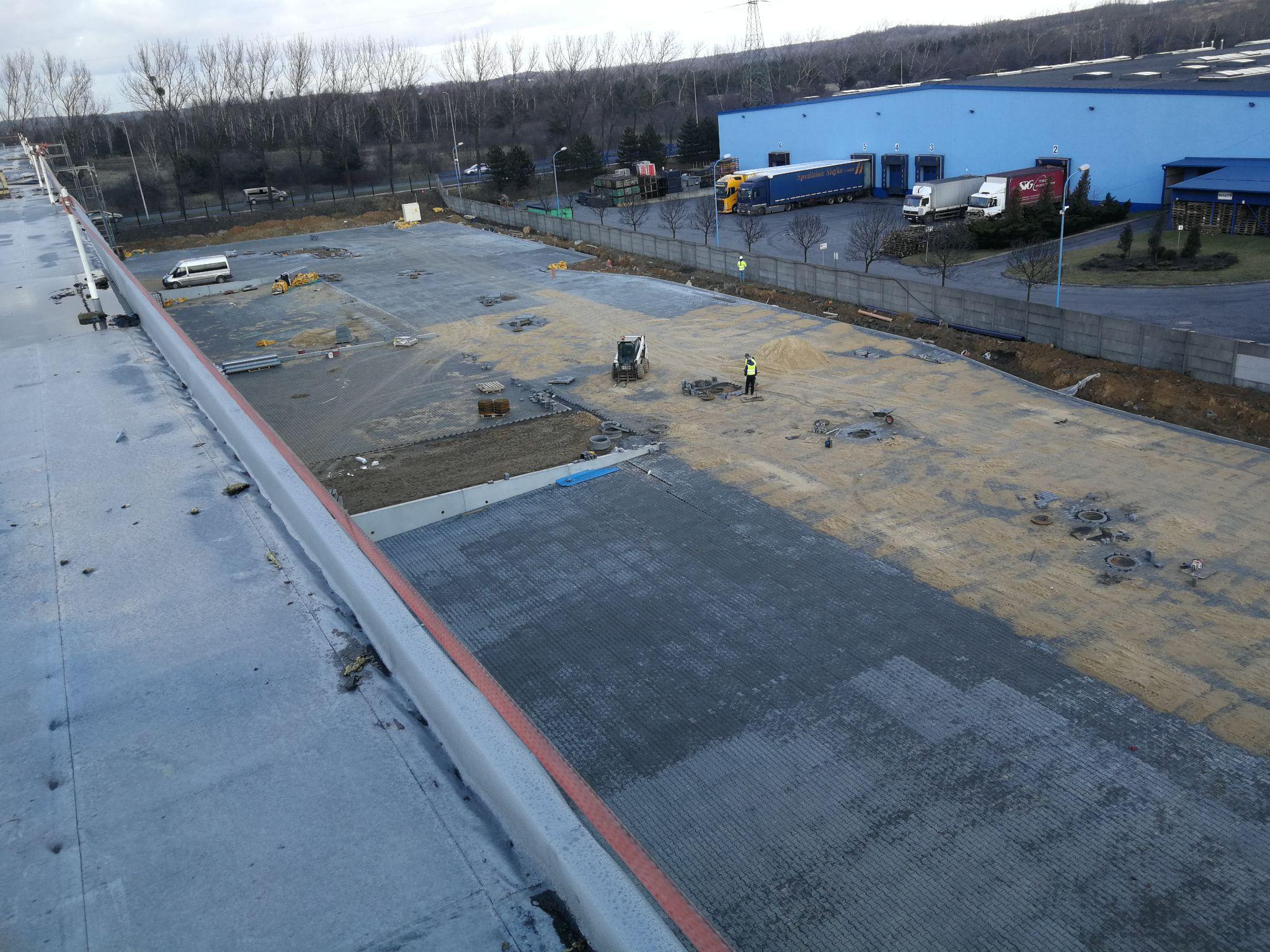 Widok z góry na plac budowy parkingu lub drogi dojazdowej. Wzdłuż lewej krawędzi widać mur lub bok dużego budynku. Duża powierzchnia jest częściowo pokryta jasnym piaskiem i częściowo siatką/geokratą, która służy jako wzmocnienie podłoża. W centrum pracują robotnicy. W tle widoczny jest inny niebieski budynek magazynowy i zaparkowane samochody dostawcze.
